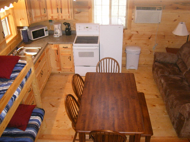 Interior of log cabin with knotty pine walls, floors and cabinets. There is a dining table, couch, bed and kitchen.