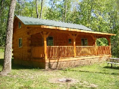 Exterior of log cabin with green roof and a full length front porch in a woodland setting.